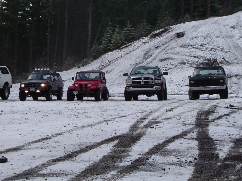 Four lifted 4x4 trucks parked in a row on a snowy gravel staging area