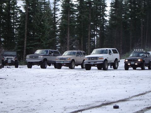 1989 Ford Bronco - A group of off-road vehicles are parked in a snowy area with a forest in the background.