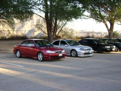 1991 Ford Taurus - Several sedans are parked in a lot under trees, with a mountainous backdrop.