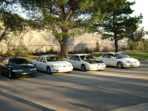 1991 Ford Taurus - Four 1991 Ford Taurus sedans are parked side-by-side in a paved lot with trees and hills in the background.