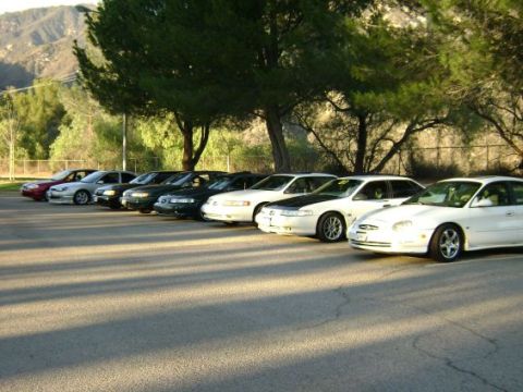 1991 Ford Taurus - A row of sedans are parked in a lot with trees and hills in the background.