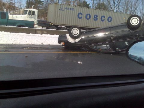 1992 Ford Taurus - A black car is overturned on the highway next to a semi-truck and trailer, with snow visible on the median.