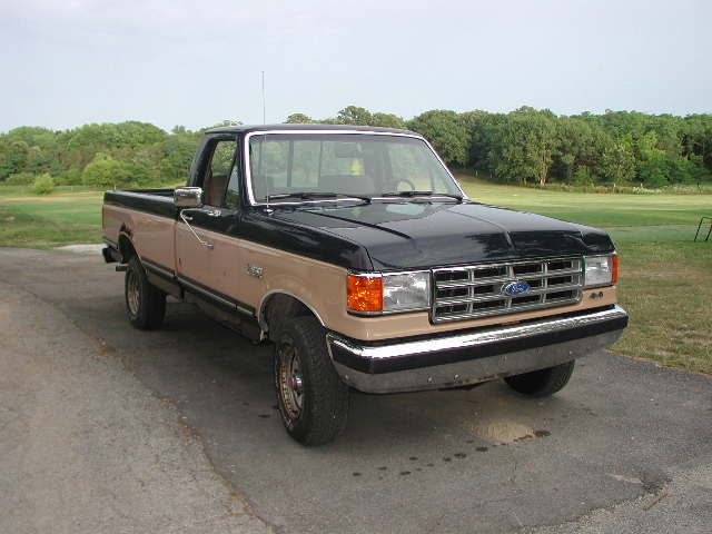Two-tone Ford F-Series pickup truck parked on an asphalt surface with trees and a field in the background