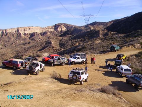 1990 Ford Bronco - A convoy of off-road vehicles gathers on a dusty trail, with a 1990 Ford Bronco among them, set against a backdrop of arid hills and a clea…