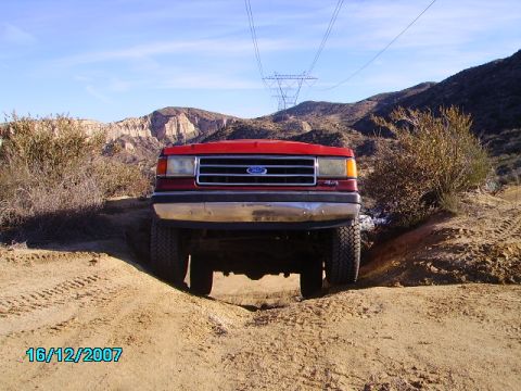 1990 Ford Bronco - A red Ford truck navigates a sandy, off-road trail under a clear blue sky.