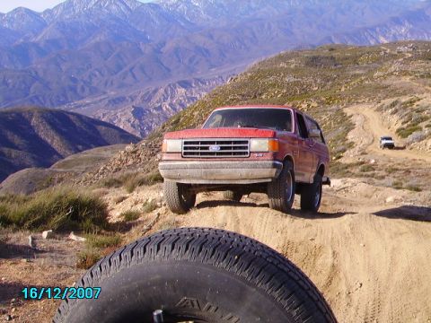 1990 Ford Bronco - A red 1990 Ford Bronco navigates a dusty mountain road, with another vehicle visible in the distance.
