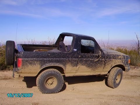 1990 Ford Bronco - 1990 Ford Bronco covered in mud after off-roading, parked on a dirt trail with a scenic overlook in the background.