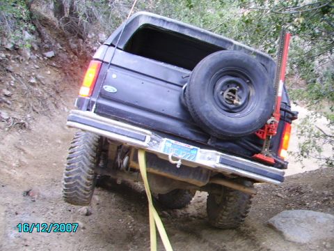 1990 Ford Bronco - 1990 Ford Bronco navigating a steep, rocky off-road trail with a high-lift jack attached.