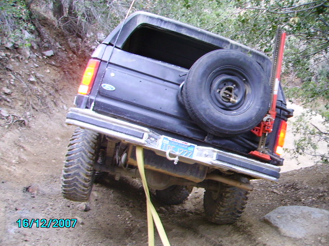 1990 Ford Bronco on a dirt trail with a high-lift jack mounted on the rear carrier