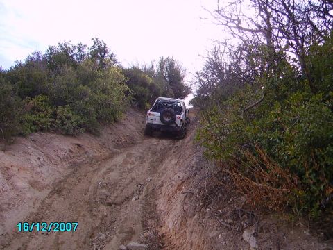 1990 Ford Bronco - A white off-road vehicle tackles a challenging dirt trail, its rear spare tire visible as it ascends the incline.