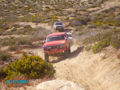 1990 Ford Bronco - Several off-road vehicles navigate a dusty trail in a desert landscape.