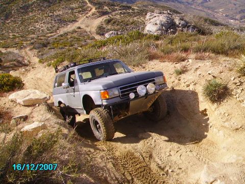 1990 Ford Bronco - This modified Ford Bronco navigates a steep, dusty off-road trail in a mountainous, arid landscape.
