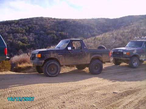1990 Ford Bronco - A muddy black 1990 Ford Bronco is parked off-road next to another vehicle.