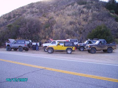 1990 Ford Bronco - Multiple off-road vehicles are parked on the side of a road near a hill.