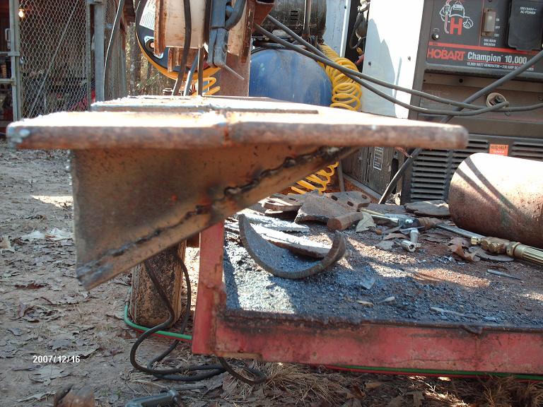Close-up of a welded metal bracket on a workbench with a Hobart welding machine in the background