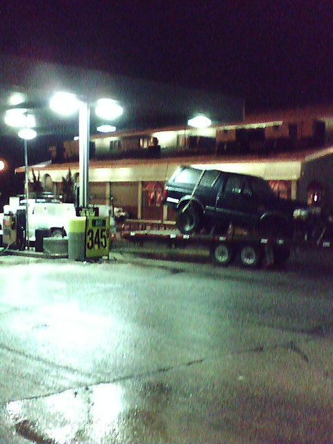 1992 Ford Bronco - A dark SUV is loaded onto a trailer at a gas station, with a price sign reading '345' visible.