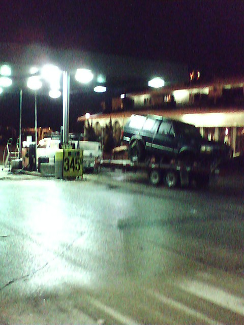1992 Ford Bronco - A dark 1992 Ford Bronco is transported on a flatbed trailer at a gas station at night.