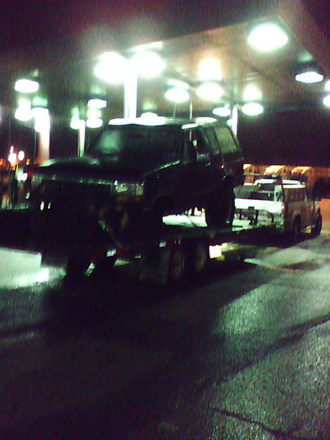 1992 Ford Bronco - A dark SUV is transported on a flatbed trailer at a gas station at night.