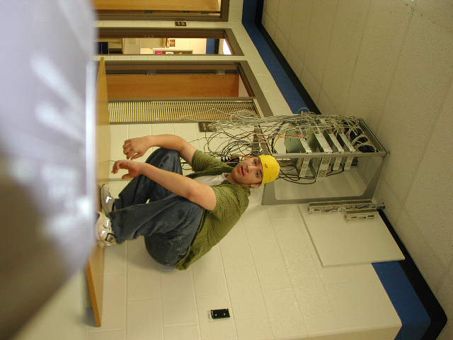 Man in yellow cap and green shirt sits on tiled floor near wiring rack