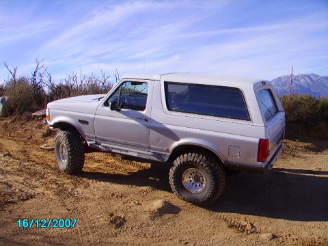 1990 Ford Bronco - A white Ford Bronco with aftermarket wheels and tires is seen off-road under a clear blue sky.