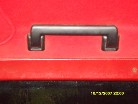 1989 Ford Bronco - Close-up of a black plastic grab handle bolted to a red painted interior panel above a window opening, with a small screw visible at right.…