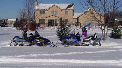 1979 Ford Bronco - Two snowmobiles are parked in a snowy yard with riders on them.
