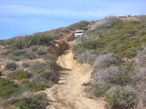 1985 Ford Bronco - A lone vehicle navigates a challenging off-road trail through arid, brush-covered hills under a clear blue sky.