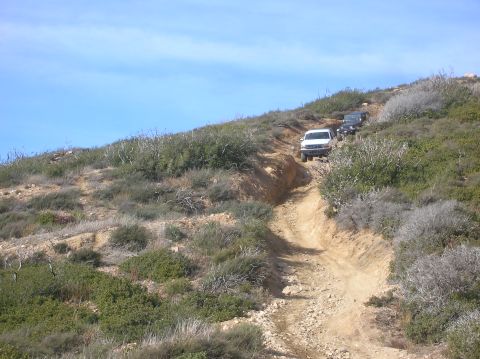 1985 Ford Bronco - Multiple off-road vehicles navigate a steep, dusty trail under a clear blue sky.