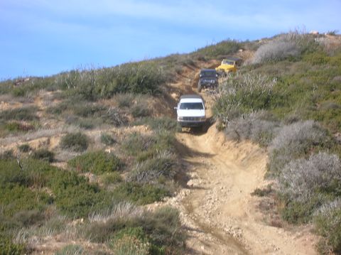 1985 Ford Bronco - Three off-road vehicles navigate a challenging dirt path on a hillside under a clear blue sky.