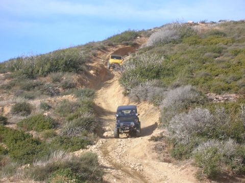1985 Ford Bronco - Two Jeeps navigate a challenging dirt trail on a sunny day.