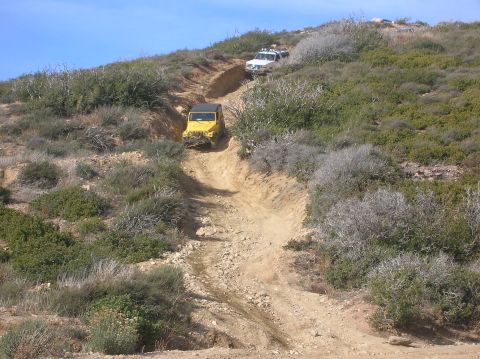 1985 Ford Bronco - Two off-road vehicles navigate a challenging dirt path through a dry, scrubby landscape under a clear blue sky.