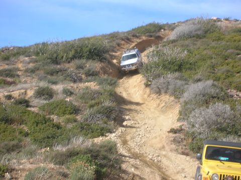 1985 Ford Bronco - Two off-road vehicles navigate a challenging dirt path on a dry, scrub-covered hillside under a clear blue sky.