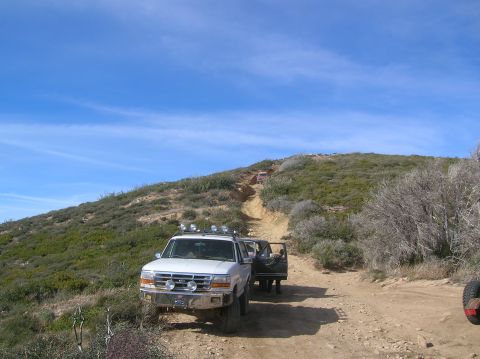 1985 Ford Bronco - A white 1995 Ford F-Series truck is parked on a dirt road, with another vehicle visible further up the path.