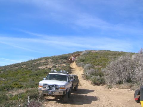 1985 Ford Bronco - Several off-road vehicles navigate a steep dirt path on a sunny day.