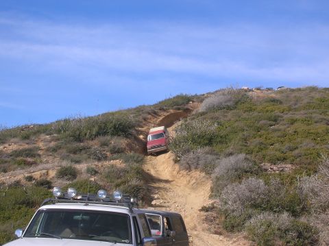 1985 Ford Bronco - Two SUVs navigate a challenging off-road trail under a clear blue sky.