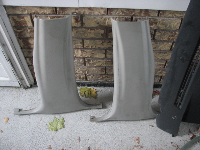 Two light gray plastic interior trim pillars and a black dashboard piece on a concrete surface with fallen leaves