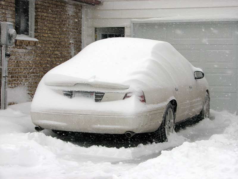 Rear view of a white 2003 Ford Crown Victoria covered in snow, parked next to a garage