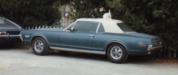 Side view of a blue coupe with white roof and a cat sitting on top, parked on gravel by a fence