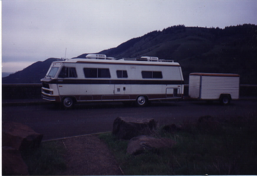 White Class A motorhome with brown stripes towing a white enclosed trailer, parked at an overlook with hills behind