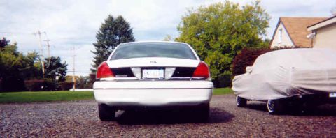 1999 Ford Crown Victoria - Rear view of a white 1998 Ford Crown Victoria parked on gravel, with another vehicle covered by a tarp to its right.