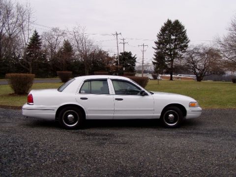 1999 Ford Crown Victoria - Side profile of a white Ford Crown Victoria sedan on a gravel surface with trees and utility poles behind it.
