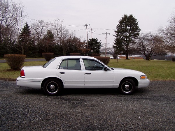White Ford Crown Victoria sedan parked on gravel, side view. Trees and overcast sky in background