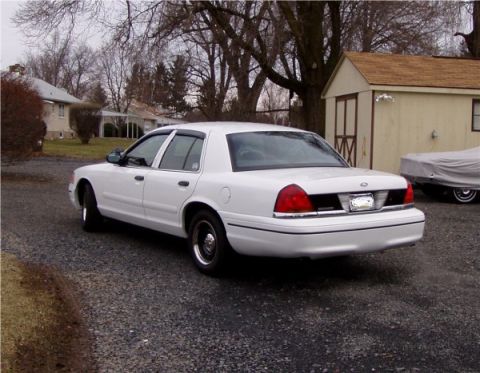 1999 Ford Crown Victoria - A white 1999 Ford Crown Victoria is parked in front of a house and shed, with another car covered in a tarp nearby.