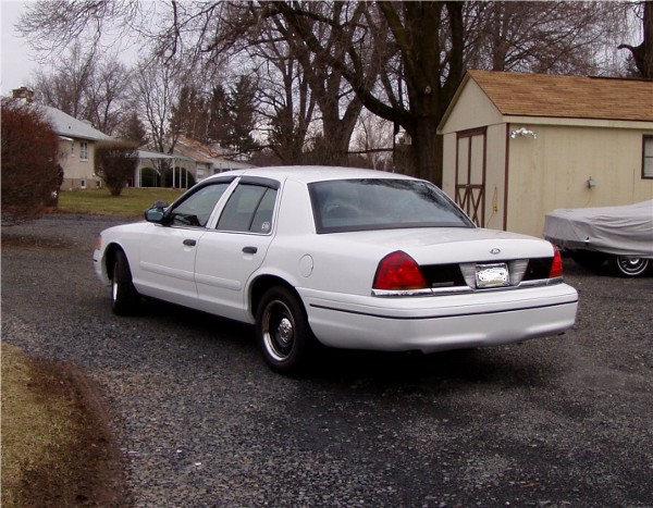 White 1999 Ford Crown Victoria parked in a driveway next to a shed. Another vehicle is covered in a tarp