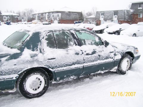 1995 Mercury Grand Marquis - A dark green sedan sits covered in snow on a winter day, with other snow-covered vehicles in the background.