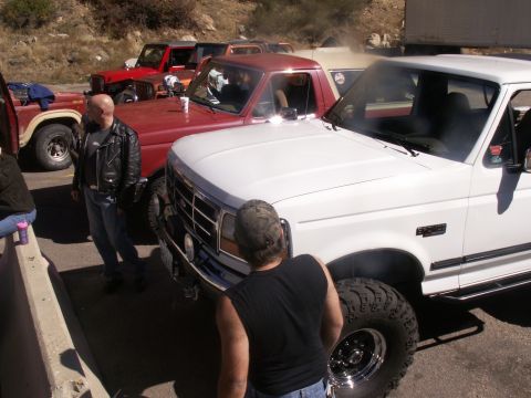 1986 Ford Bronco - Several trucks are parked on a roadside, with a white Ford F-Series truck in the foreground emitting smoke.
