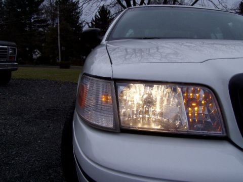 1999 Ford Crown Victoria - A white sedan's front corner is shown, with the headlight on, and another vehicle in the background.