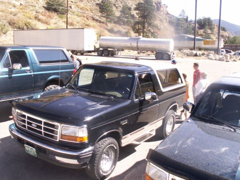 1986 Ford Bronco - A black Ford Bronco with a white top is parked next to another vehicle, with a semi-trailer truck in the background.