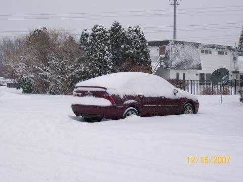 1995 Mercury Grand Marquis - A maroon sedan is covered in snow on a cloudy day, with a house and trees in the background.