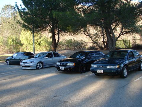 1992 Ford Taurus - A lineup of four sedans, including a dark green 1992 Ford Taurus, parked under trees in a lot.